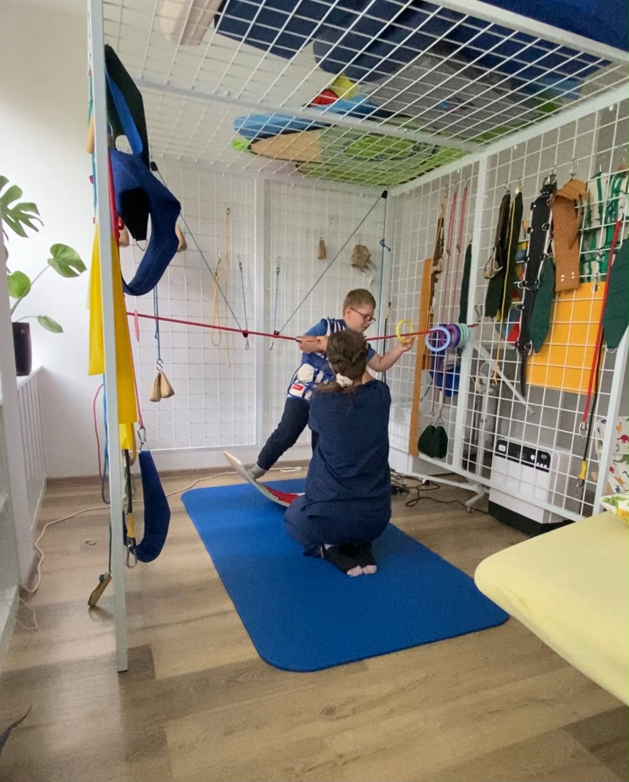 Leuško drží tyč s krúžkami v terapeutickej klietke, terapeutka kľačí vedľa neho a pomáha mu