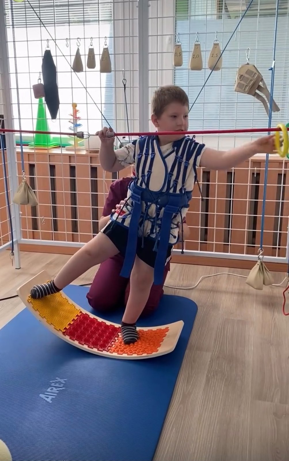 Leuško v terapeutickom postroji balansuje na kolísavej doske a naťahuje sa po tyči s krúžkom
